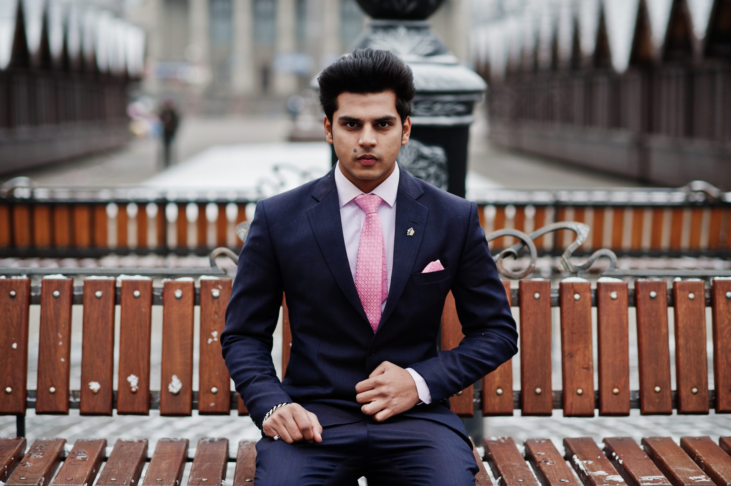 Elegant indian macho man model on suit and pink tie posed on winter day and sitting on bench.